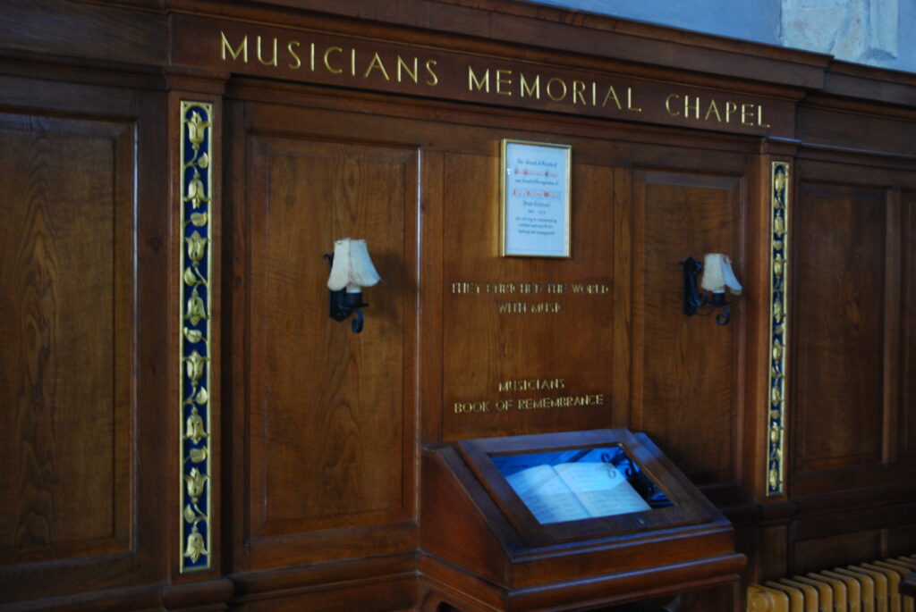 A wood panelled wall, with the text Musicians' Memorial Chapel carved above. A display case with a glass frontage, displaying an open book of remembrance